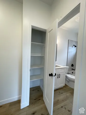 Built-in storage closet featuring tiered shelving, wood-finish flooring, white Shaker-style cabinetry with a rectangular sink, and tile-surround shower with matte black fixtures