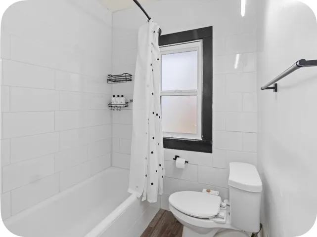 Full bathtub with white subway tile surround, a window with dark trim, and wood-finish flooring