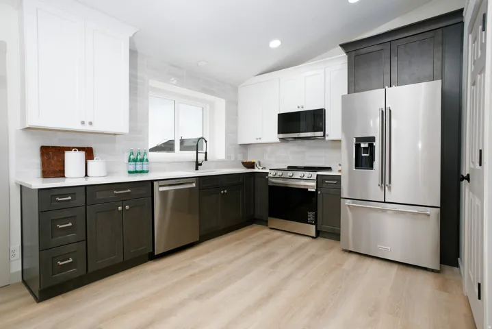 Two-tone cabinetry with white upper and dark lower units, stainless steel appliances, white countertops, subway tile backsplash, and light wood-finish flooring