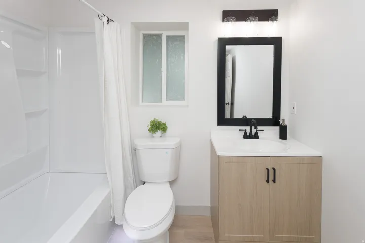 White bathtub with shower, light wood-finish vanity with white countertop, black-framed mirror, and matte black faucet