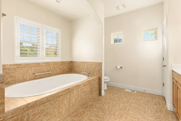 Soaking tub with tile surround, white plantation shutters, neutral tile flooring, and light wood-finish cabinetry