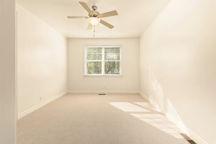 Neutral-toned carpeting in a spacious room with a window, white trim, and a ceiling fan