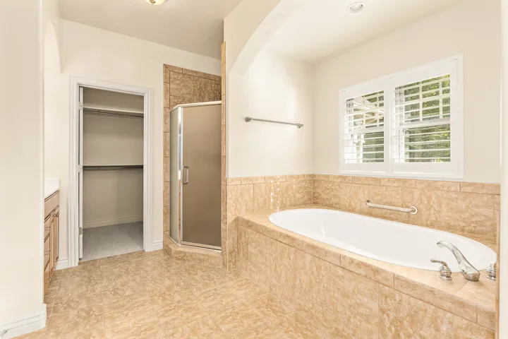 Built-in soaking tub with tile surround, walk-in shower with frosted glass enclosure, and white plantation shutters
