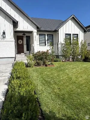 White facade with vertical siding accents, dark roof, dark-framed windows, and an established lawn with mature shrubs