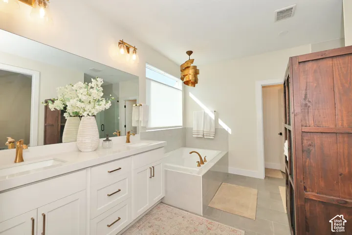 Bathroom featuring a dual-sink vanity with a solid-surface countertop, gold-tone hardware, a deep soaking tub with a tiled surround, and large-format neutral floor tile