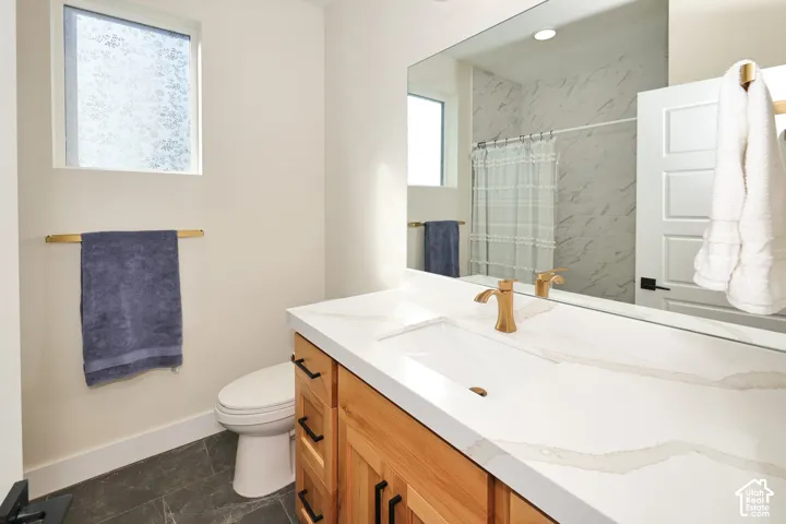 Bathroom featuring a wood-finish vanity with a stone-composite countertop, undermount sink, brass-tone faucet, and full-width vanity mirror, complemented by dark stone-tile flooring and a shower surround with decorative wall tile