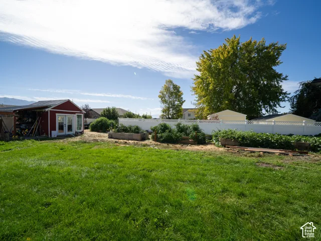 Expansive grassy area with a red storage shed featuring double glass doors, raised garden beds, and a white privacy fence