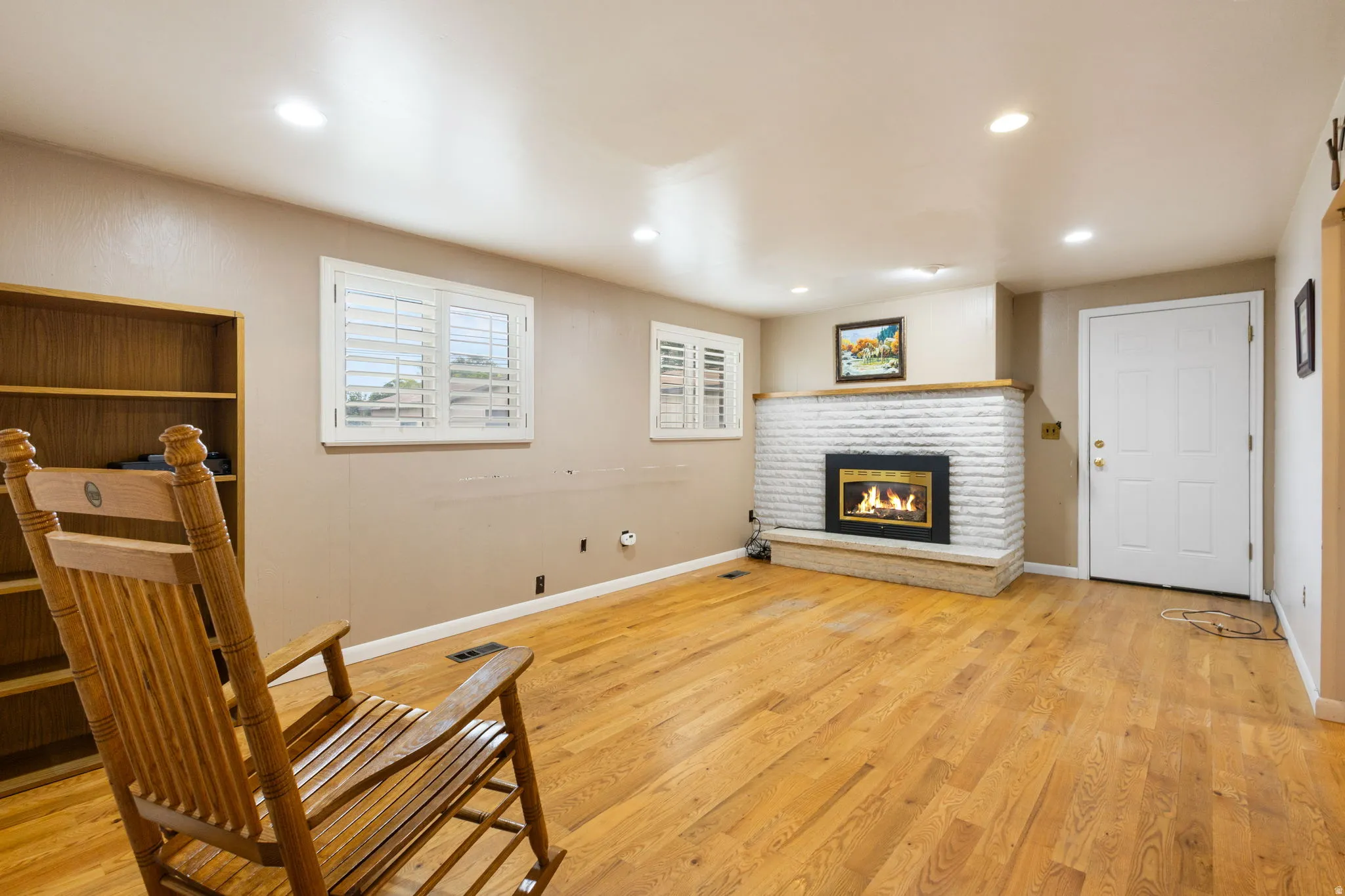 Hardwood flooring throughout, white brick fireplace with hearth, built-in shelving, recessed lighting, and windows with white plantation shutters