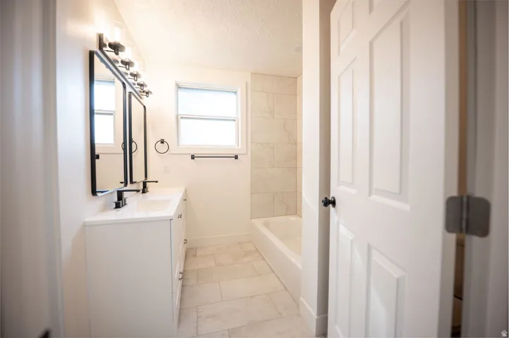 Dual vanity with white cabinetry and integrated sinks, matte black hardware, tiled flooring, and a tiled bath surround