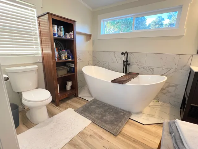 Freestanding soaking tub with a floor-mounted matte black faucet, wall-mounted rectangular tiles, and wood-finish flooring