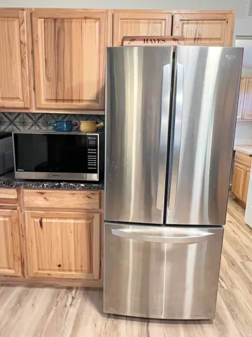 Natural wood-finish cabinetry, stainless steel French door refrigerator, dark speckled countertops, decorative tile backsplash, and light wood-finish flooring