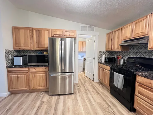 Kitchen featuring wood-finish cabinetry, stainless steel refrigerator, black range, patterned tile backsplash, and light wood-finish flooring