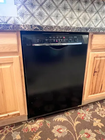 Built-in black dishwasher with a stainless steel handle, complemented by light wood cabinetry, a patterned tile backsplash, and a dark, patterned countertop