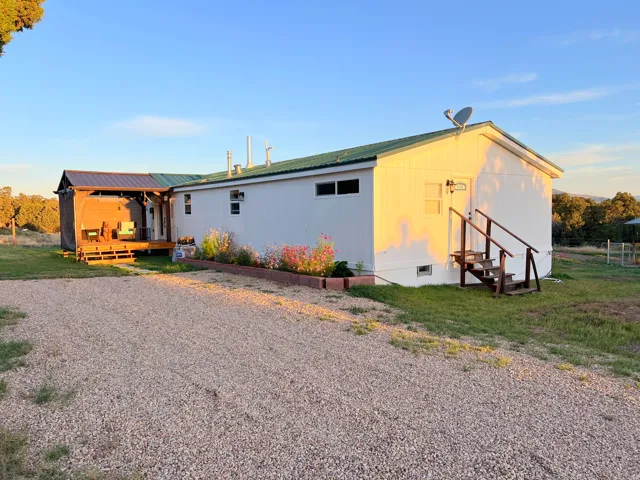White exterior with a green metal roof, a gravel driveway, a covered wood deck, and a raised garden bed with flowering plants