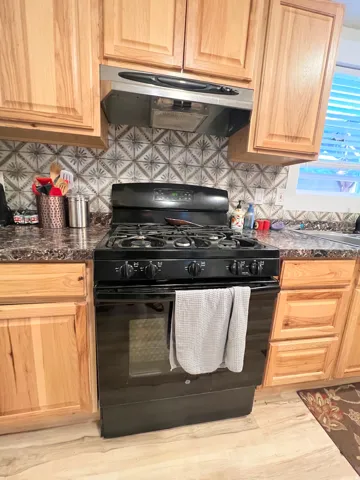 Black gas range with an overhead vent hood, wood cabinetry, patterned backsplash, and wood-finish flooring