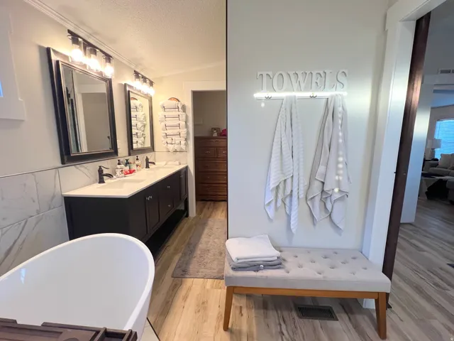 Freestanding soaking tub, dual vanity with undermount sinks, wall-mounted light fixtures, wood-finish flooring, and crown molding