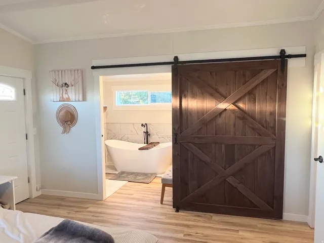 Wood-finish flooring, solid wood barn door with black hardware, freestanding soaking tub, tiled bath surround, and transom window