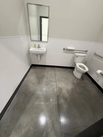 Wall-mounted sink with chrome faucet, wall-mounted mirror, toilet, and concrete flooring