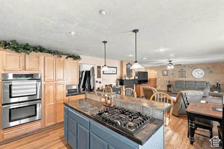 Kitchen featuring light wood cabinetry, stainless steel double wall ovens, wood-finish flooring, and an island with a built-in gas cooktop and pendant lighting