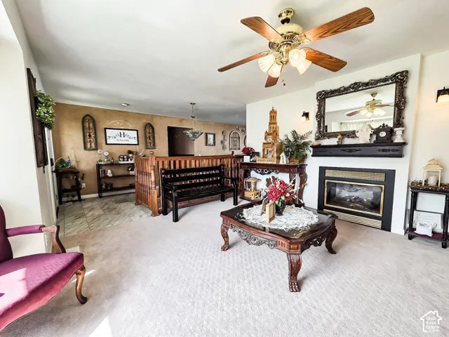 Living area featuring a fireplace with a black mantel, carpet flooring, a ceiling fan with light fixture, and a partial wood railing