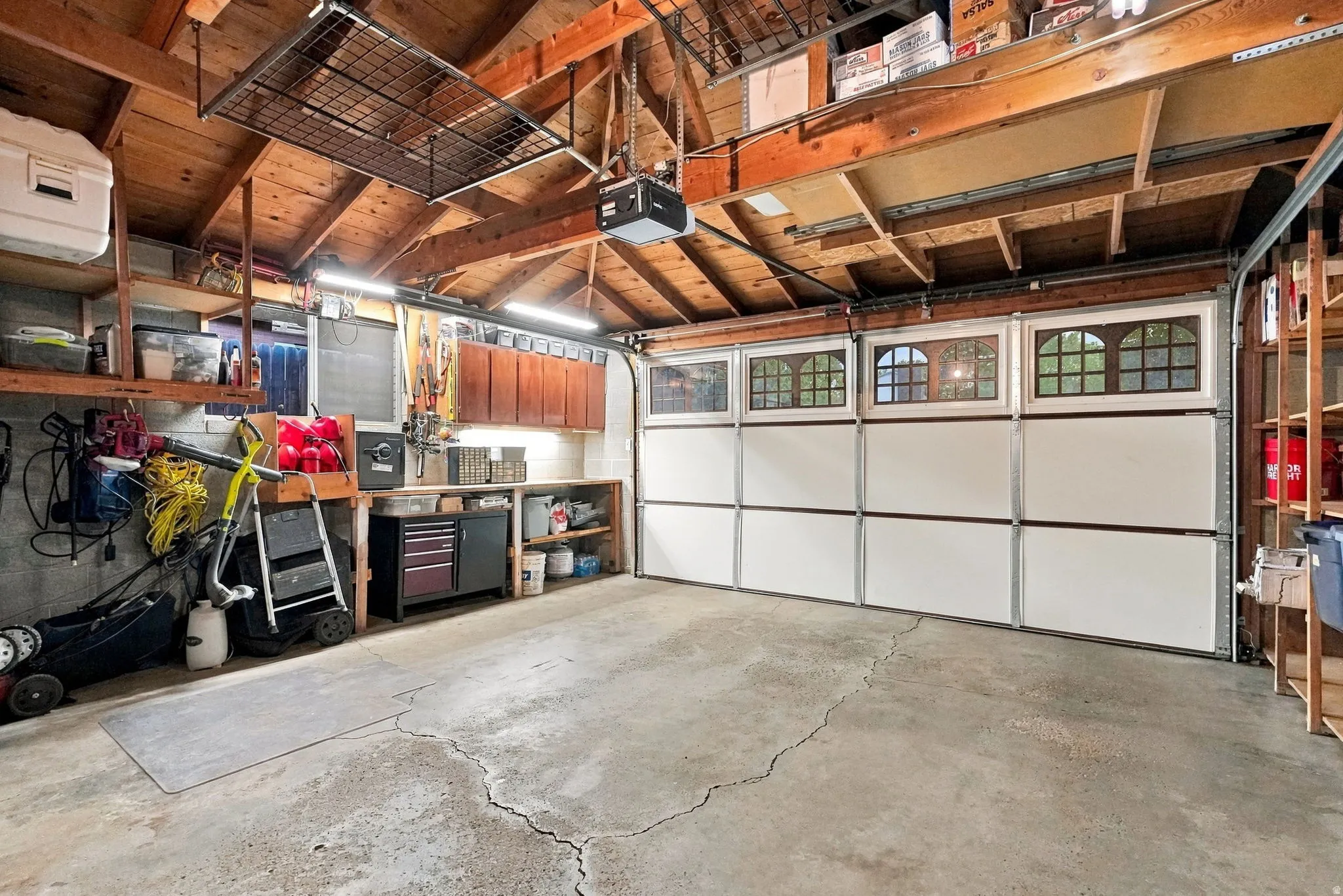 Concrete flooring garage with an exposed wood beam ceiling, suspended overhead storage, and built-in wall shelving