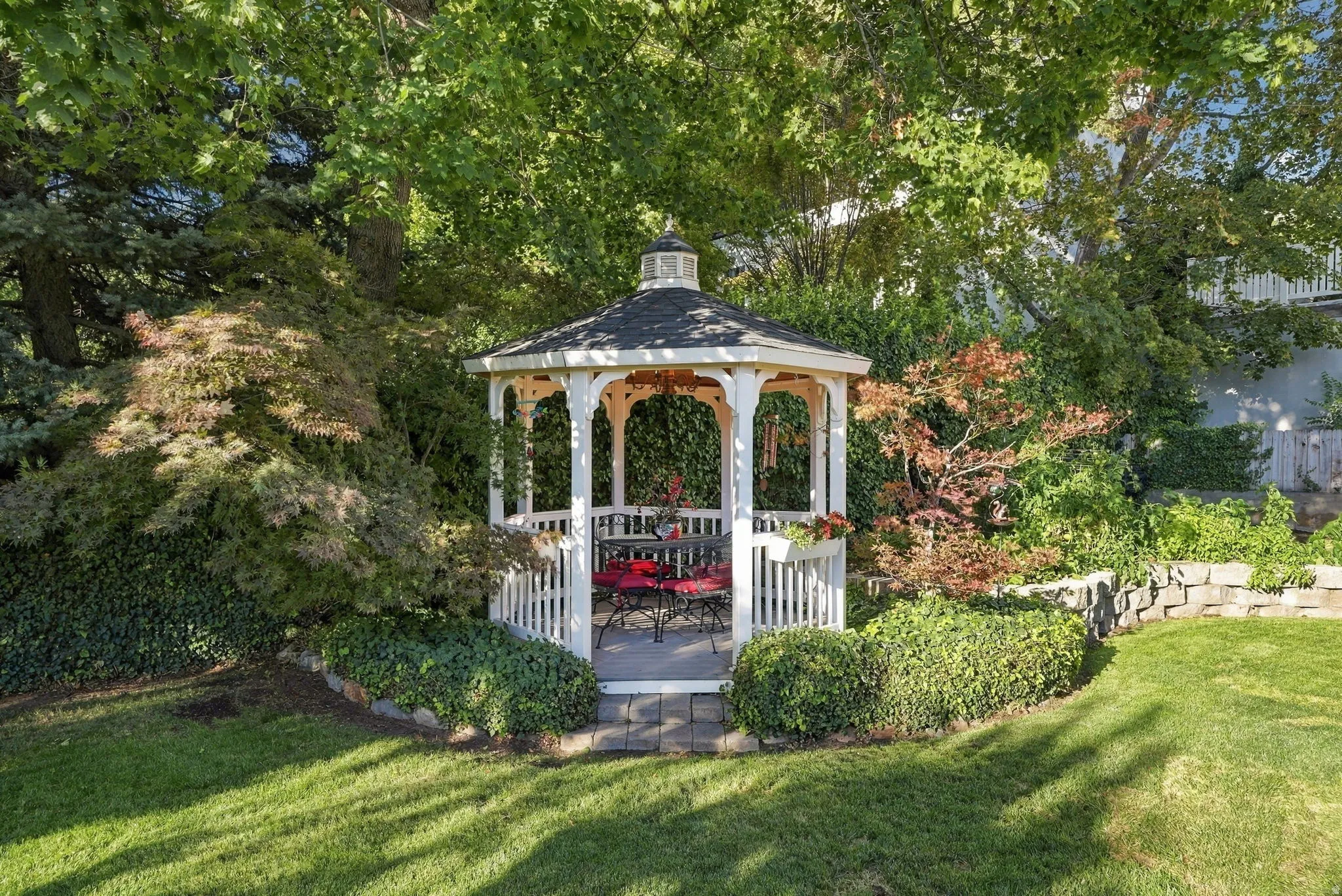 White wood gazebo with a dark shingle roof, surrounded by mature trees and shrubs