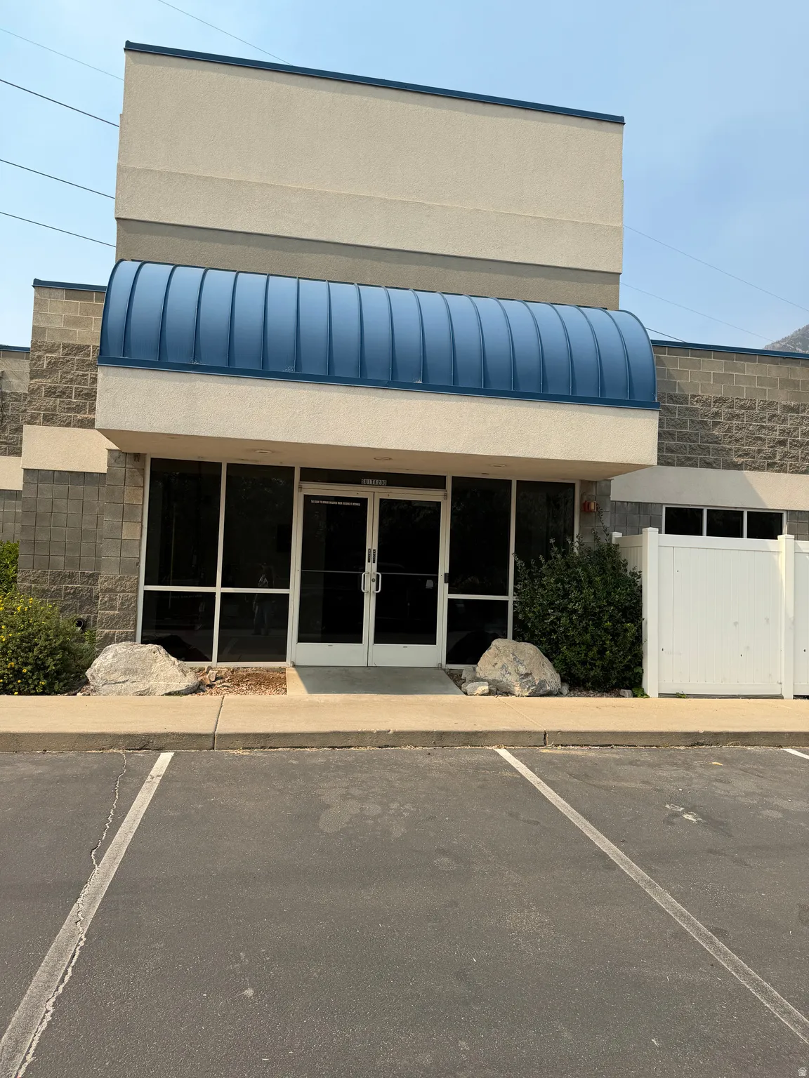 Stucco and block exterior building featuring a prominent blue curved awning, dual glass entry doors, and a paved parking area