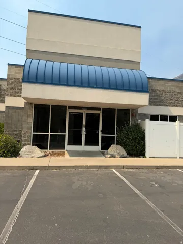 Stucco and block exterior building featuring a prominent blue curved awning, dual glass entry doors, and a paved parking area