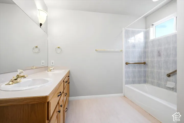 Double vanity with gold-tone fixtures and integrated sinks, wood-finish cabinetry, full-width mirror, and tub with tiled surround