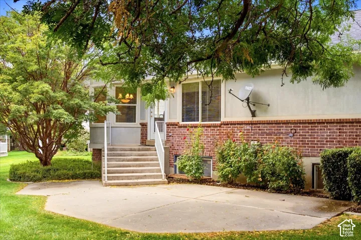 Rear exterior featuring a concrete patio, brick and stucco siding, exterior lighting, concrete steps with white railings, and mature trees