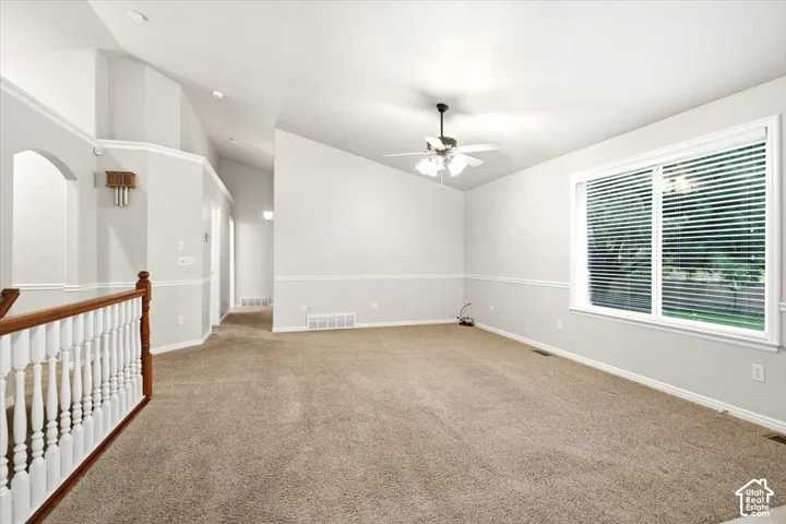 Spacious carpeted room featuring a large window, vaulted ceilings, a ceiling fan, and architectural archway details