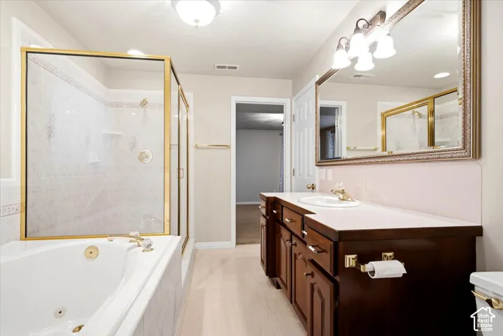 Double vanity with wood cabinetry, large vanity mirror with decorative frame, light-toned wood-finish flooring, and a jetted soaking tub with tile surround