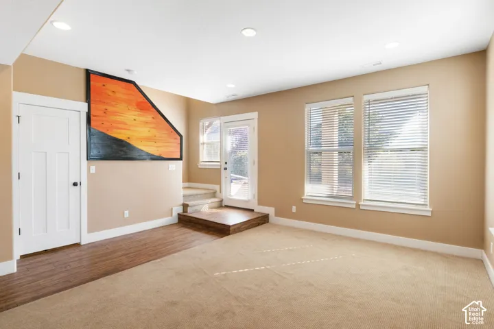 Recessed ceiling lighting, wood-finish flooring, two large windows with blinds, built-in carpeted stairs, and a white paneled door