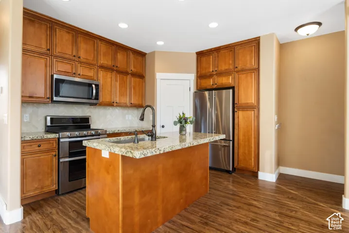 Kitchen featuring wood-finish cabinetry, stainless steel appliances, a center island with a sink, stone countertops, and wood-finish flooring