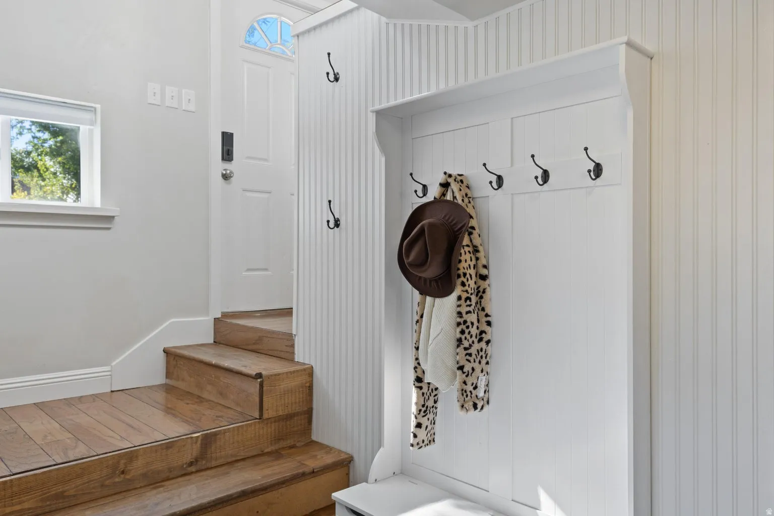 Entryway featuring wood-finish stairs, a white paneled door with an arched transom, a built-in white coat rack with black hooks, and white beadboard wall paneling