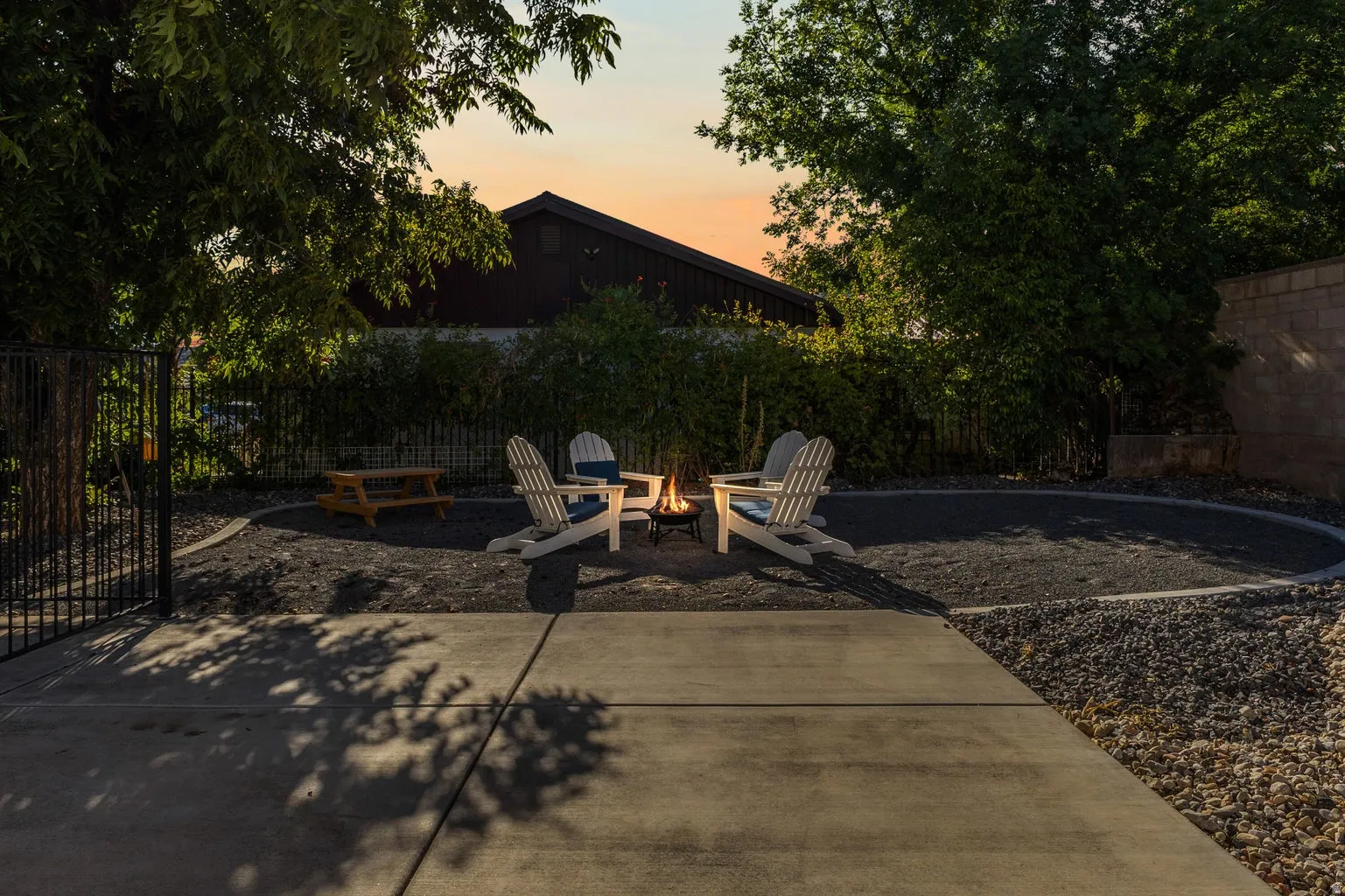 Concrete patio with a circular fire pit area, surrounded by dark gravel and mature trees, featuring a black metal fence
