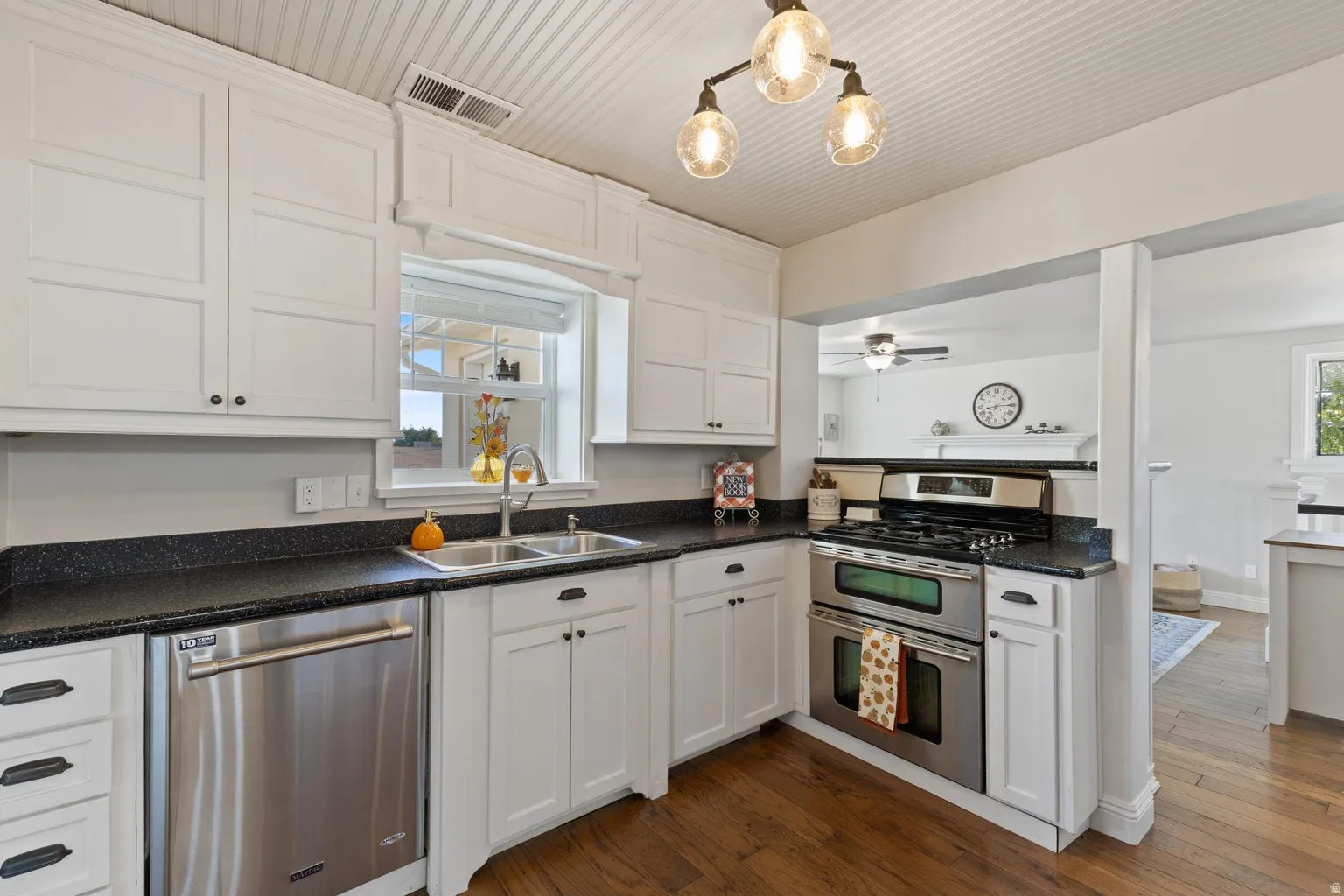 White shaker-style cabinetry with dark hardware, stainless steel appliances, dark countertops, a double basin sink, and wood-finish flooring