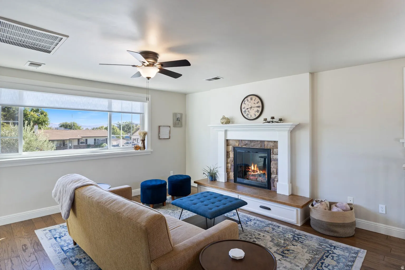 Wood-finish flooring, a ceiling fan with integrated lighting, a large picture window, and a fireplace with a stone surround and wood mantel