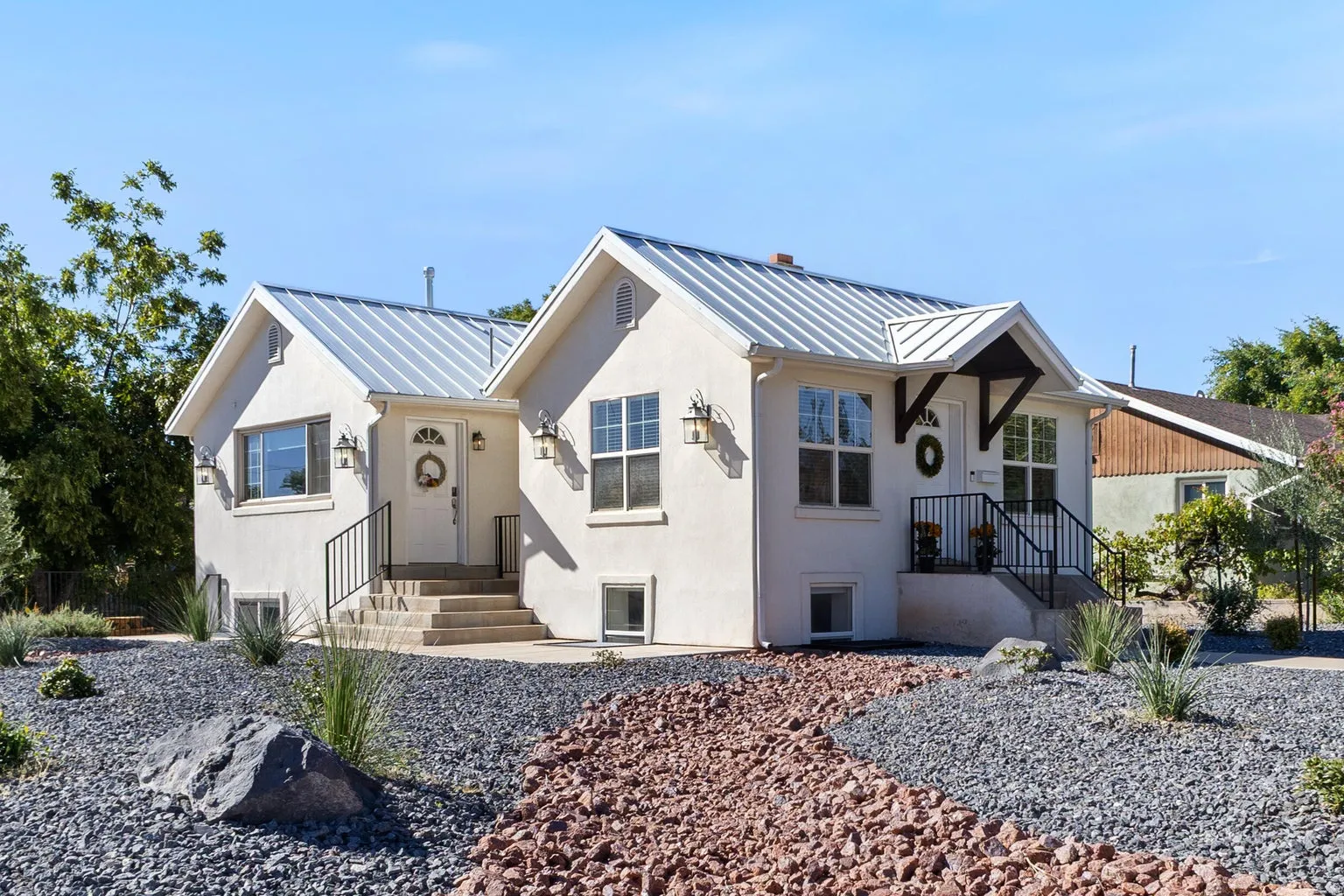 Stucco exterior featuring dual entryways with metal roofs, exterior sconce lighting, and a xeriscaped front yard with decorative rock and plantings