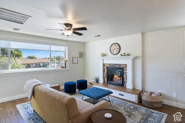 Wood-finish flooring, a ceiling fan with integrated lighting, a large picture window, and a fireplace with a stone surround and wood mantel