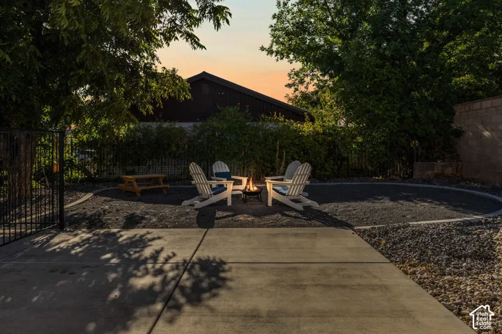 Concrete patio with a circular fire pit area, surrounded by dark gravel and mature trees, featuring a black metal fence