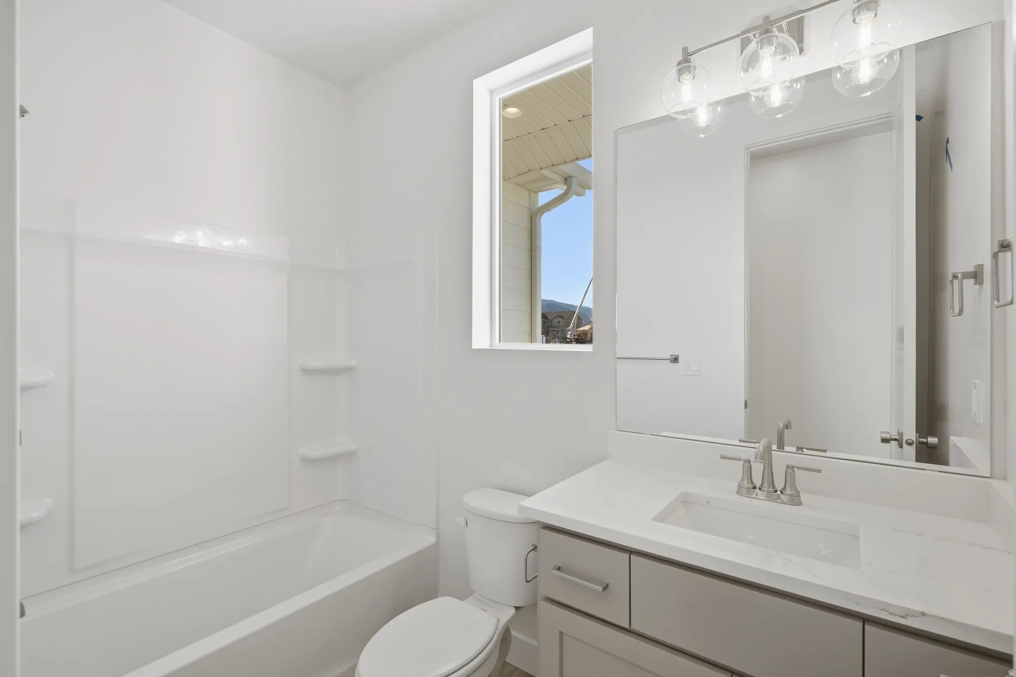White bathtub with integrated shelving, a rectangular window, a vanity with a rectangular undermount sink, a stone-finish countertop, and a three-light fixture