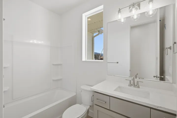 White bathtub with integrated shelving, a rectangular window, a vanity with a rectangular undermount sink, a stone-finish countertop, and a three-light fixture