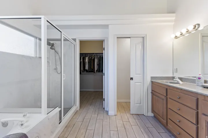 Dual vanity with integrated sinks, a glass-enclosed shower, built-in bathtub, wood-finish flooring, and a walk-in closet