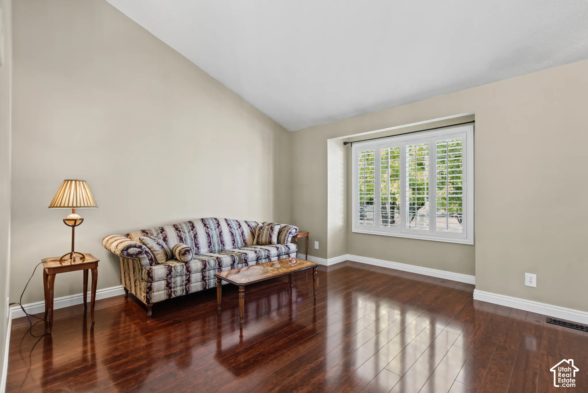 Rich wood-finish flooring, vaulted ceiling, and window with white plantation shutters
