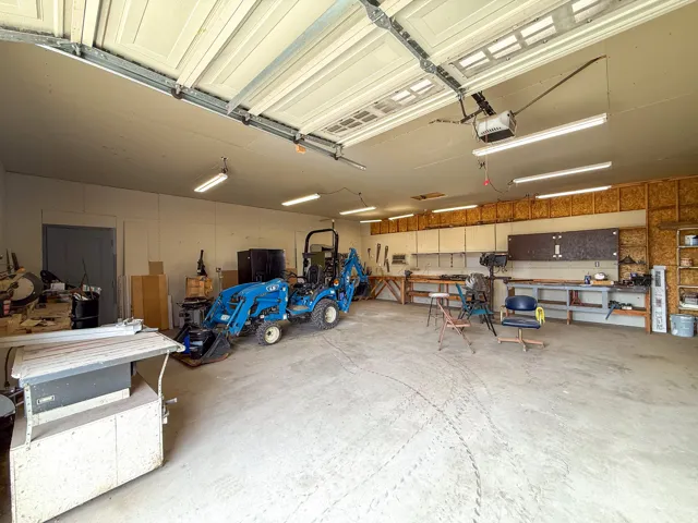 Spacious attached garage with multiple workbenches, built-in cabinetry, overhead lighting, and a concrete floor