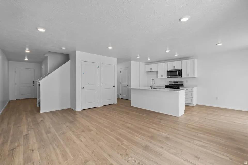 Open-concept living area with wood-finish flooring, recessed lighting, and a kitchen featuring white cabinetry, a center island with sink, and stainless steel appliances