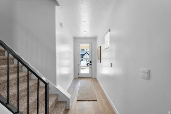 Light wood-finish flooring, recessed ceiling lighting, a white painted interior, a white door with clear glass panels, and a black wrought iron staircase railing