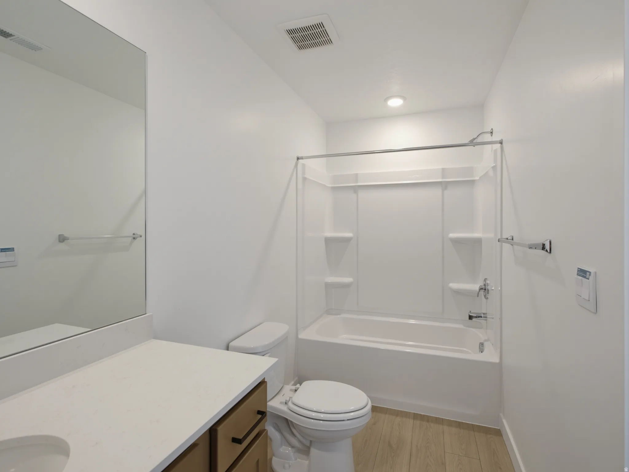 Recessed lighting illuminating a white bathtub with a shower surround, wood-finish flooring, a white toilet, and a vanity with a light-toned countertop and wood-finish cabinetry