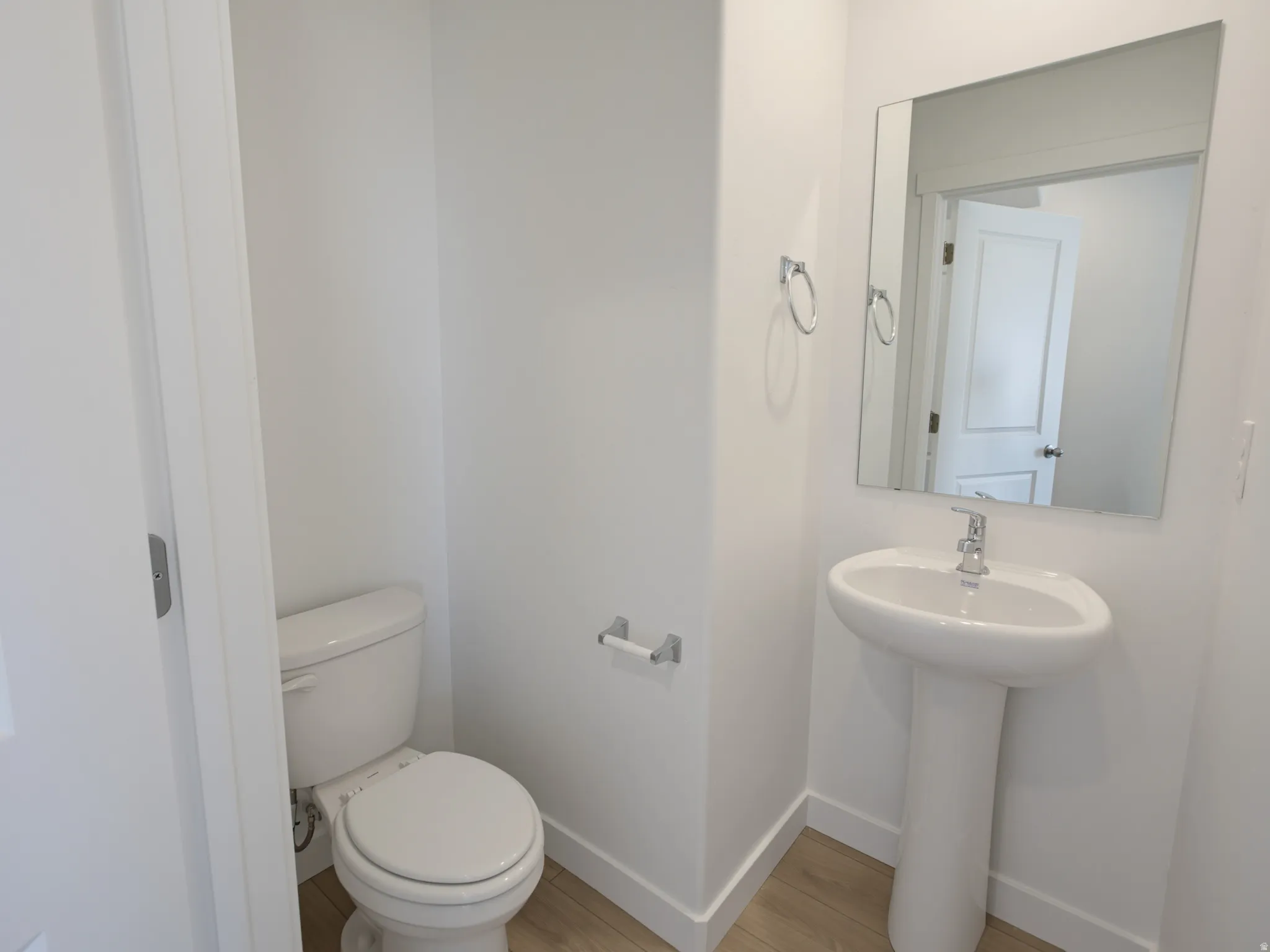 Pedestal sink with chrome faucet, wall-mounted mirror, white toilet, and wood-finish flooring